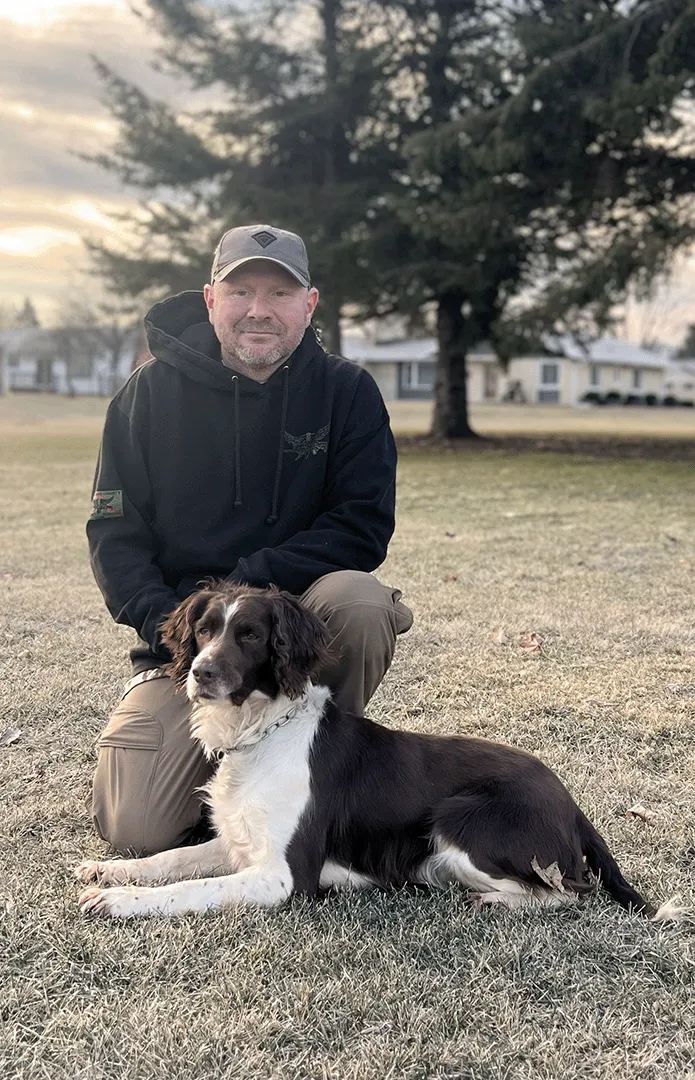 Brandon kneeling in a field with one of his family dogs laying in front of him.