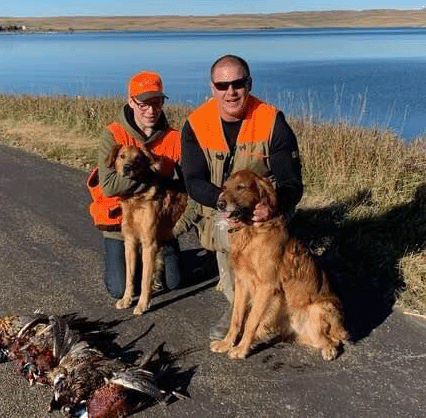 Two men in bird hunting atire kneeling and petting two golden retrievers.